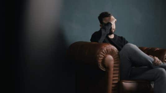 man sittng in brown sofa holding his forehead