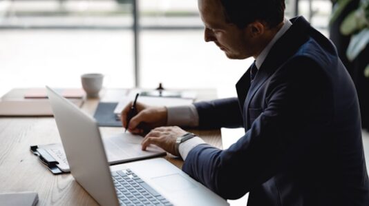 a man writing some notes with laptop open on the table
